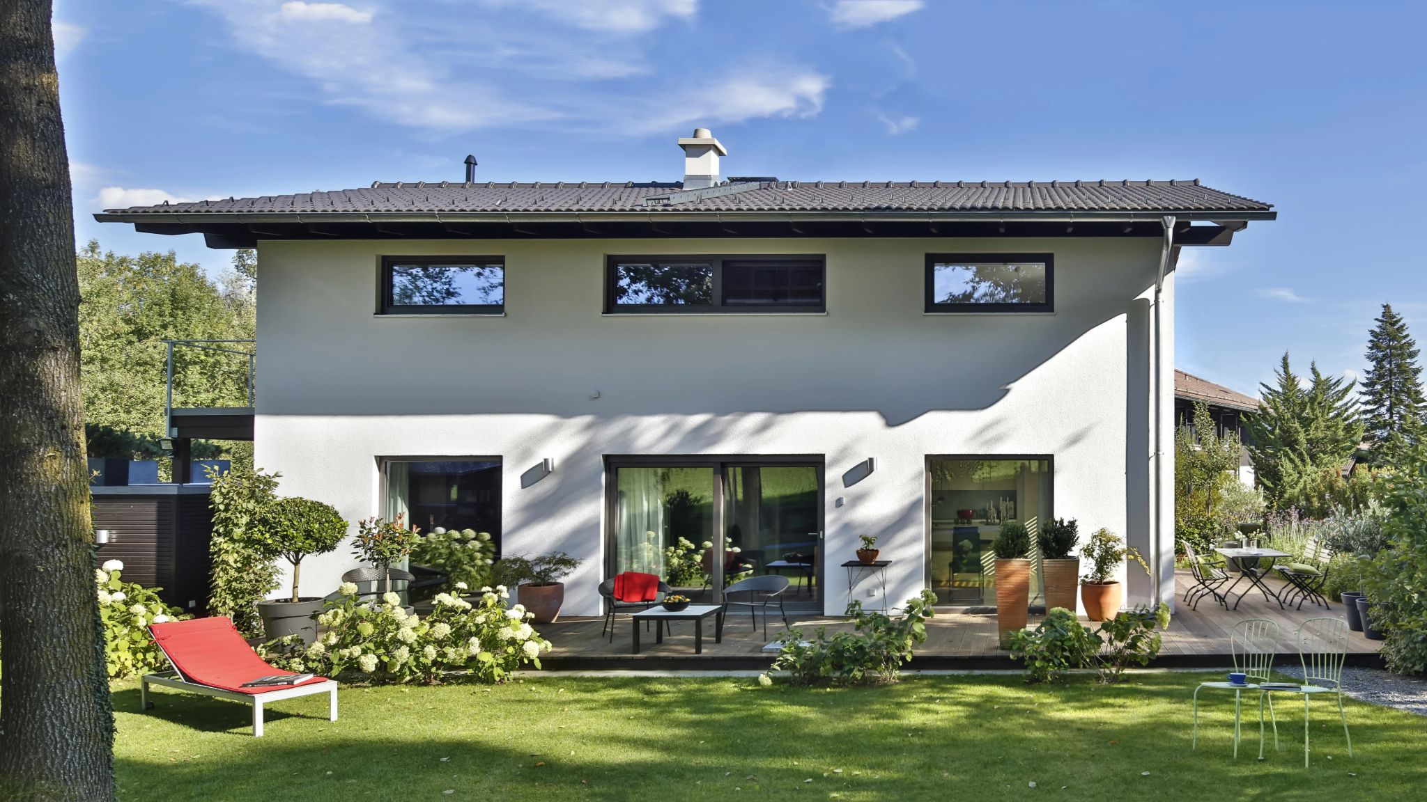 Modernes Einfamilienhaus mit Terrasse, Pool, Gartenmöbeln und üppiger Bepflanzung an einem sonnigen Tag.
