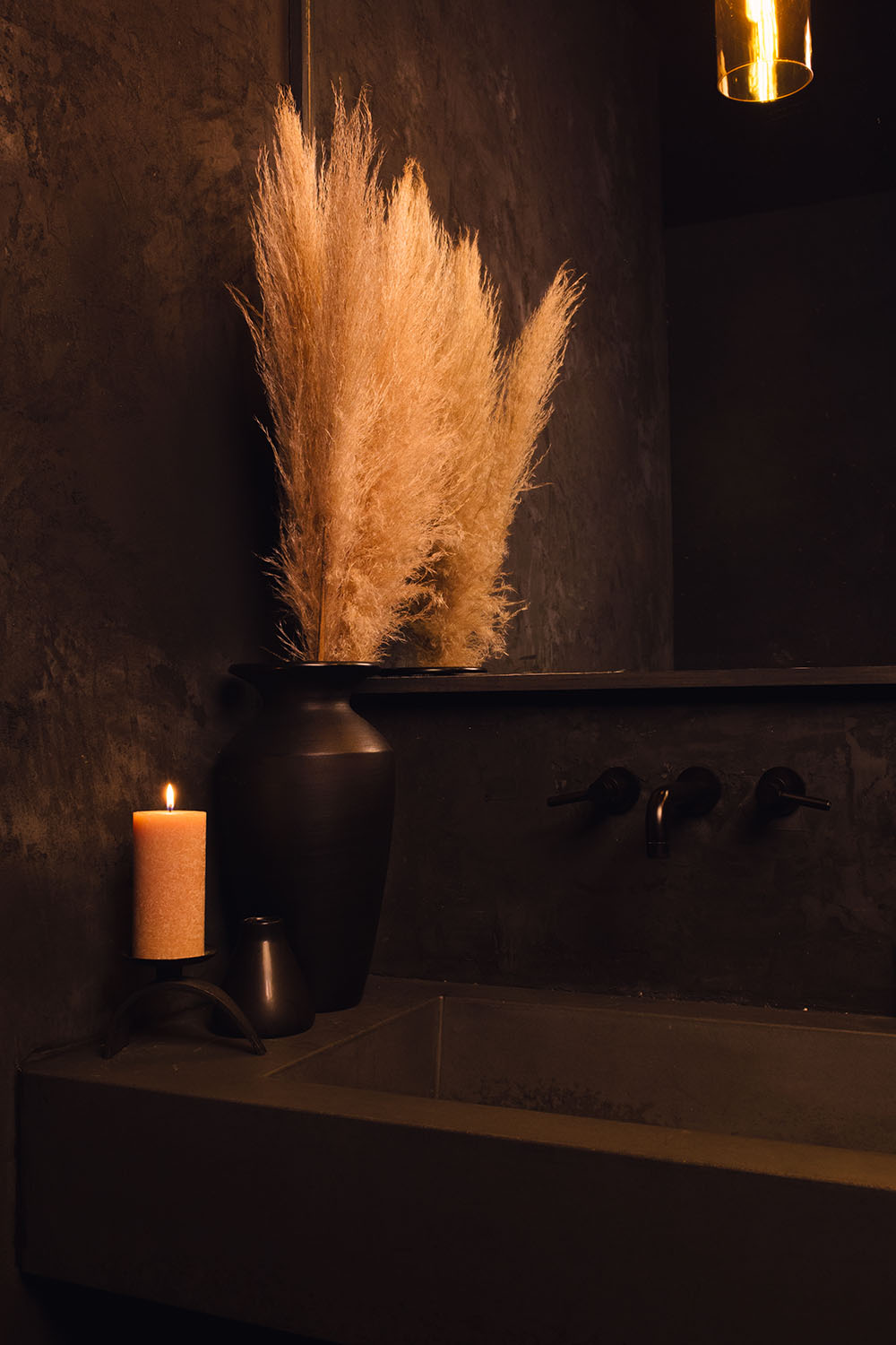 A dried yellow plant resembling wheat sits in stone dark stone vase next to a lit yellow candle of the same hue. The candle and the vase sit on the sink in the washroom of Particle Studio in Seattle, WA.