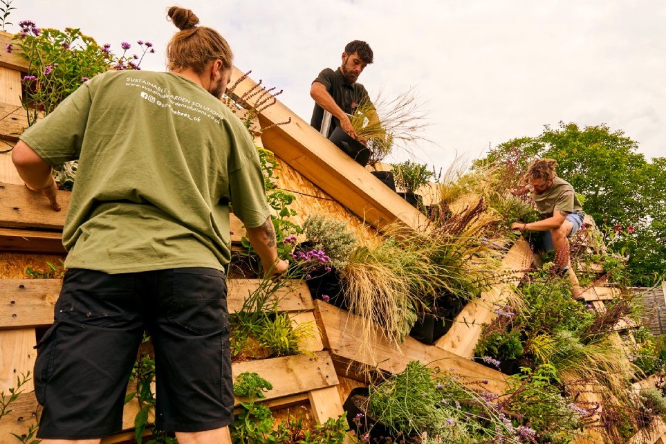 Three individuals work together on a wooden structure, arranging logs amidst a natural outdoor setting.