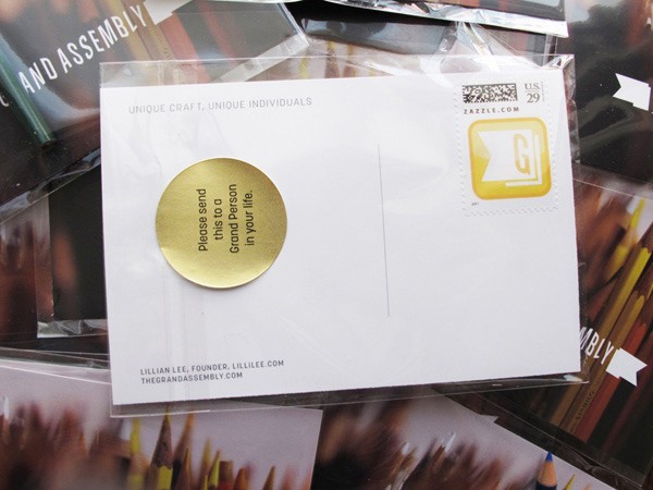 A white envelope with a gold circular sticker reading “Please send this to a Grand Person in your life.” rests on a clear plastic bag containing other envelopes with printed text and images of colored pencils. The envelope also displays “UNIQUE CRAFT, UNIQUE INDIVIDUALS” at the top and “ZAZZLE.COM” in the upper right corner. The sticker is affixed to the center of the envelope.