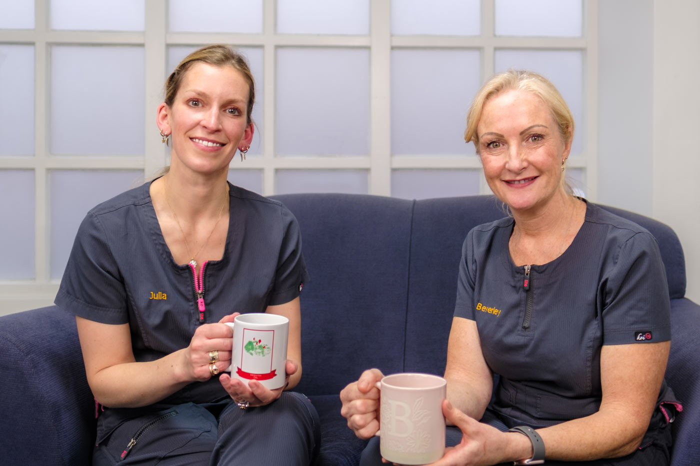 Two women, Julia and Beverley, both wearing dark blue dental scrubs embroidered with their names and "Bell House Dental Practice," are sitting side-by-side on a blue sofa. They are smiling and holding mugs, enjoying a hot drink. Julia holds a Christmas-themed mug, while Beverley holds a pink mug with a large letter 'B' on it.