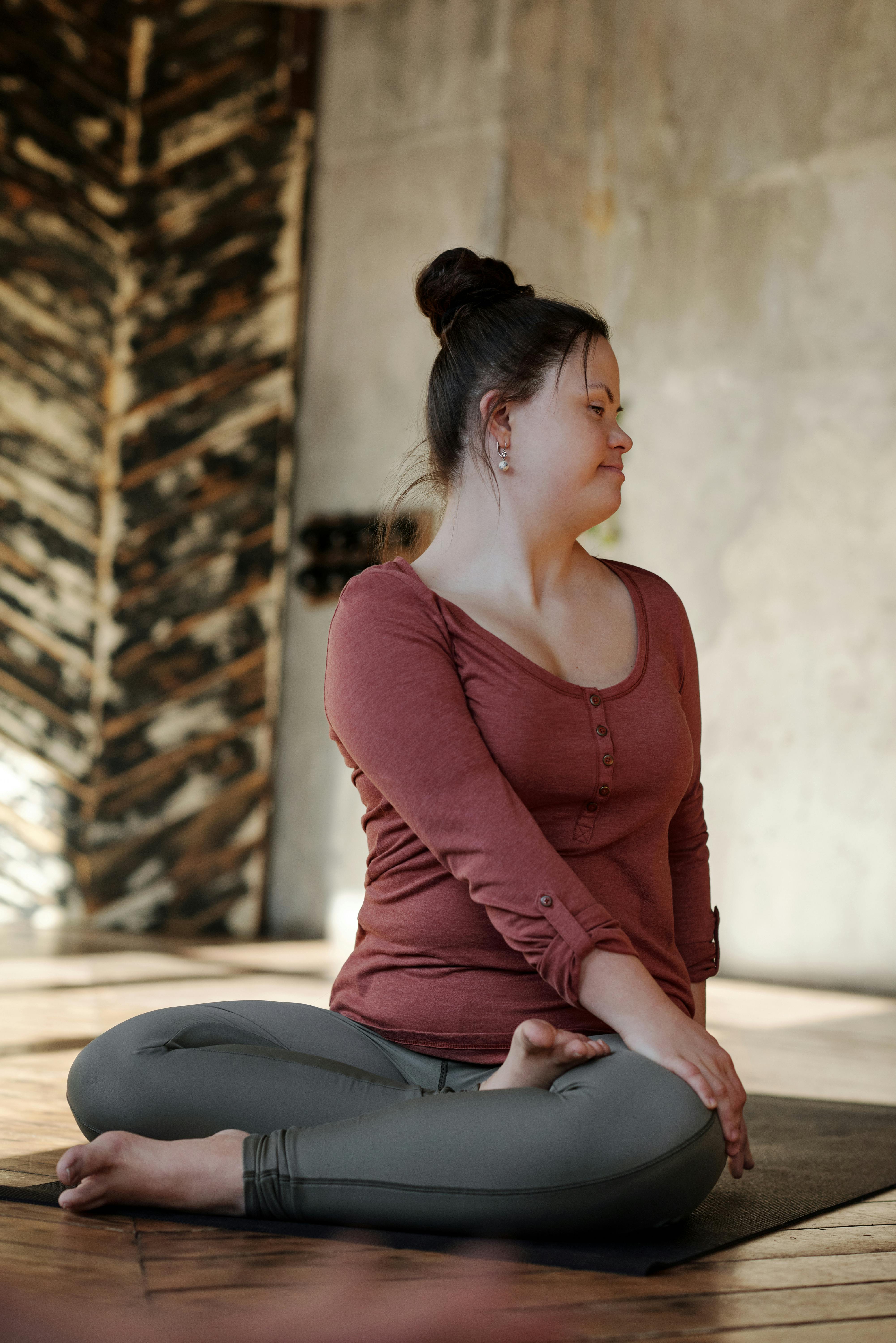 A woman with her hair in a bun sits cross-legged on a yoga mat, twisting her torso. She's wearing a rust-colored top and grey leggings, exuding calm focus.