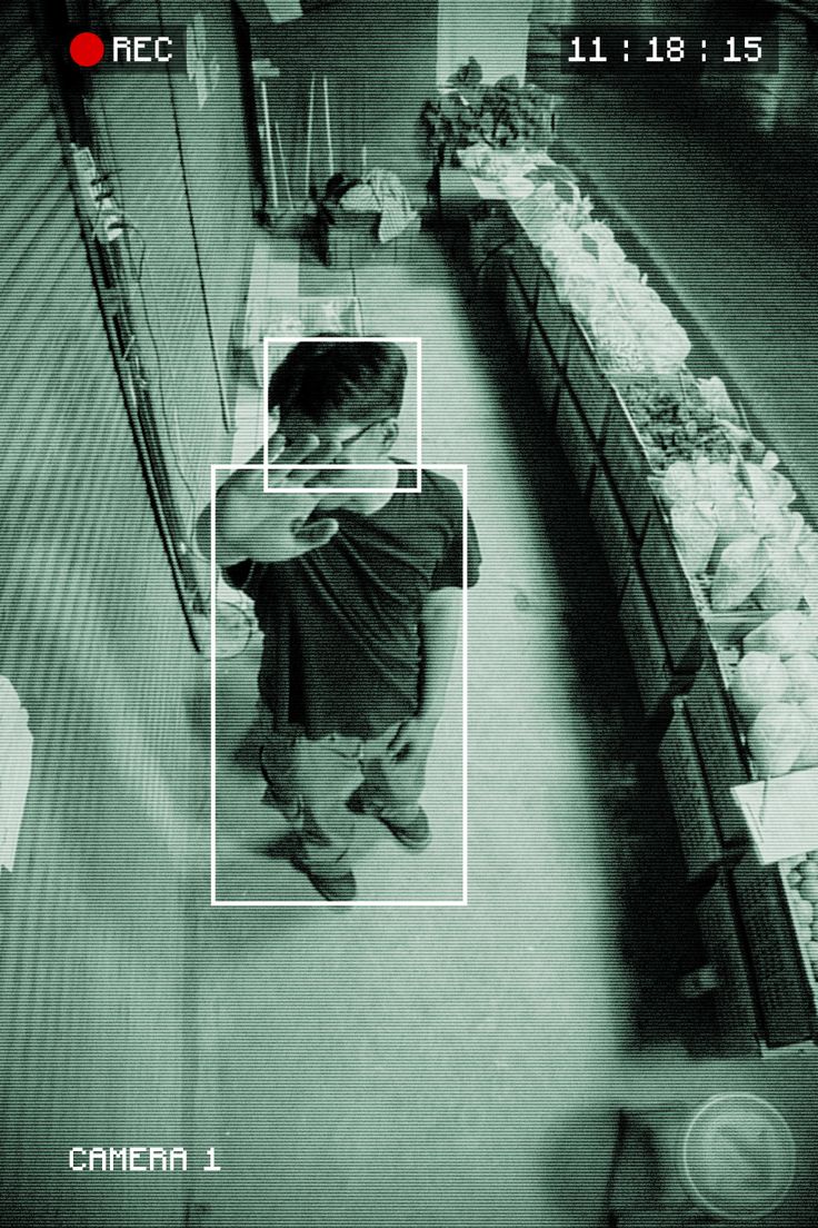A person stands at an indoor market stall lined with fresh produce, captured in a grainy, surveillance camera-style black-and-white photograph, emphasizing a sense of candid observation and everyday life.