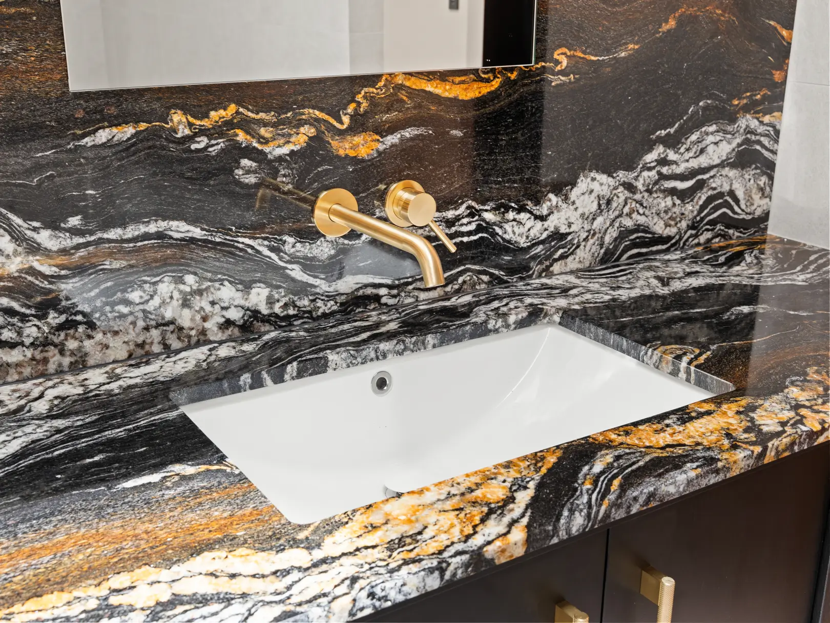 Detail shot of a high-end custom vanity featuring a dramatic black, gold, and white veined stone benchtop and splashback, an undermount white sink, and minimalist brass wall-mounted tapware.