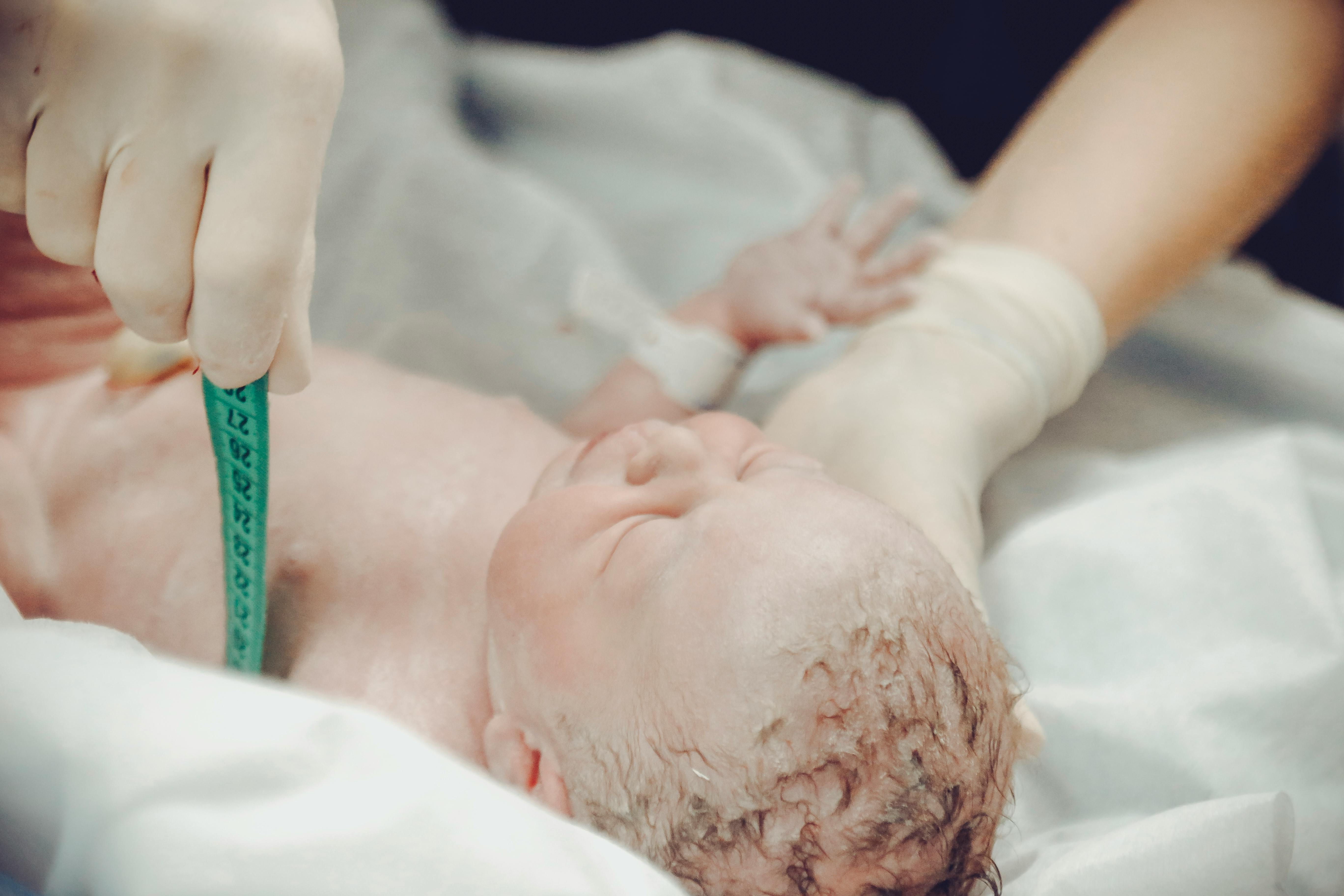A close up of a newborn being measured with a measuring tape. It is lying on a white sheet being supported by gloved hands.