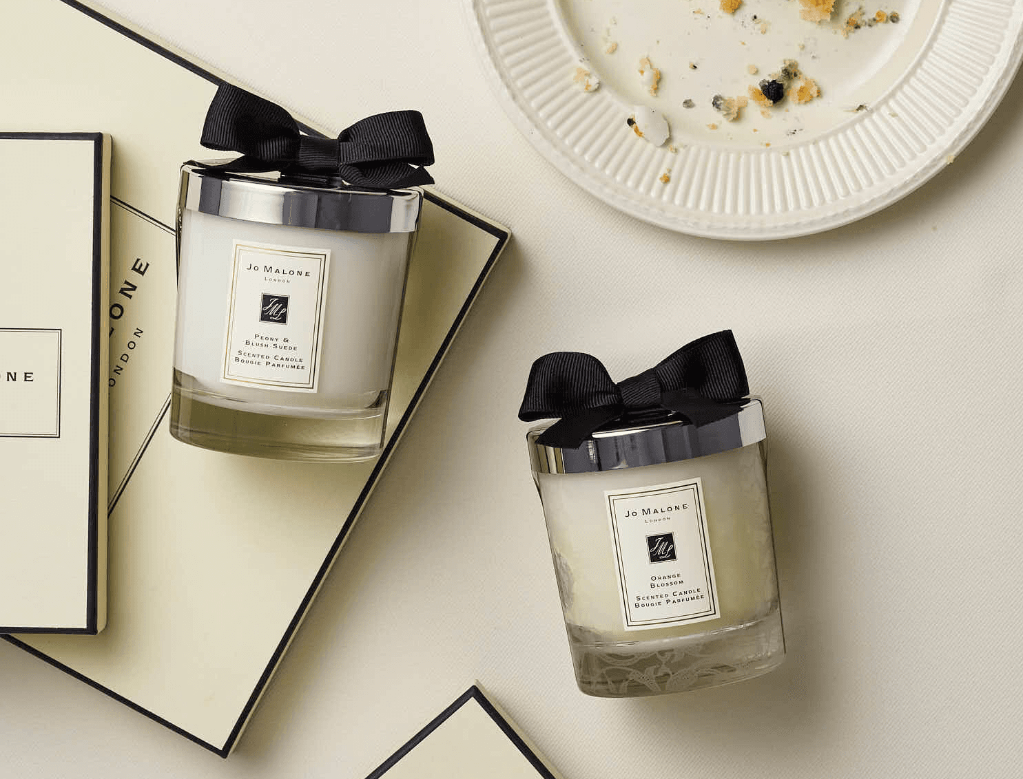 Elegant scented candles with black ribbon, placed on Jo Malone branded packaging, next to a plate with crumbs.