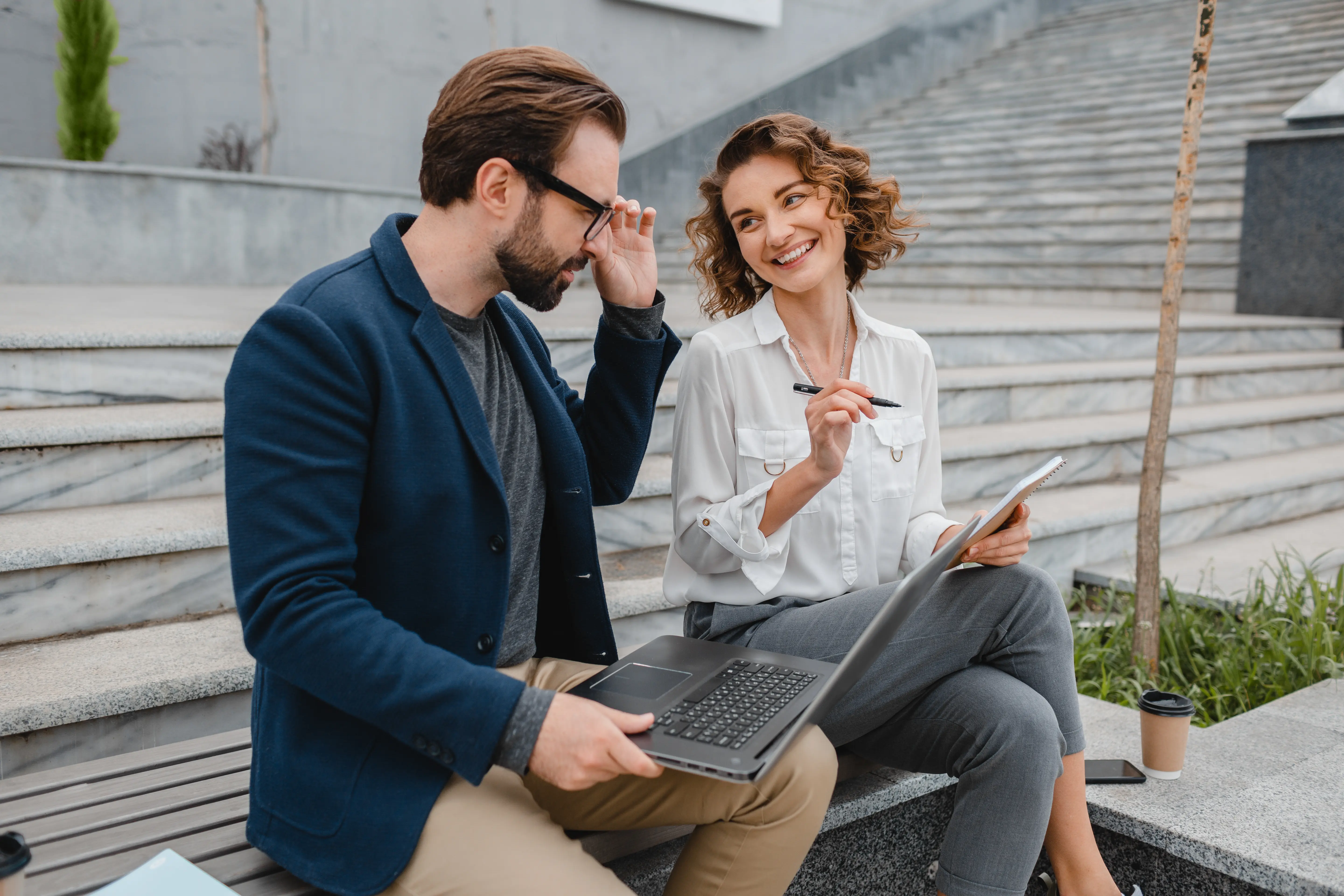 Smartly dressed man and woman sitting down on stairs. The man is holding a laptop and the woman is holding a pen and a notebook