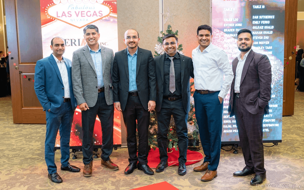 Six men from the SteriMax team pose together in professional attire at a Las Vegas-themed holiday party, standing between a festive welcome sign and a digital seating chart.