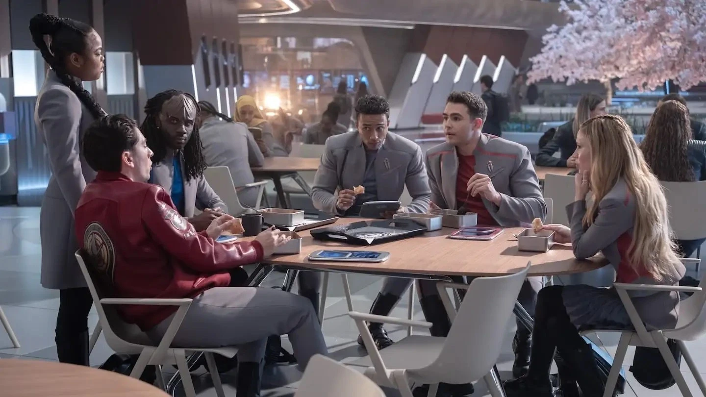 A diverse group of young people in futuristic uniforms sit around a table in a sleek, modern space with a blooming pink tree in the background, engaging in conversation and eating light snacks.