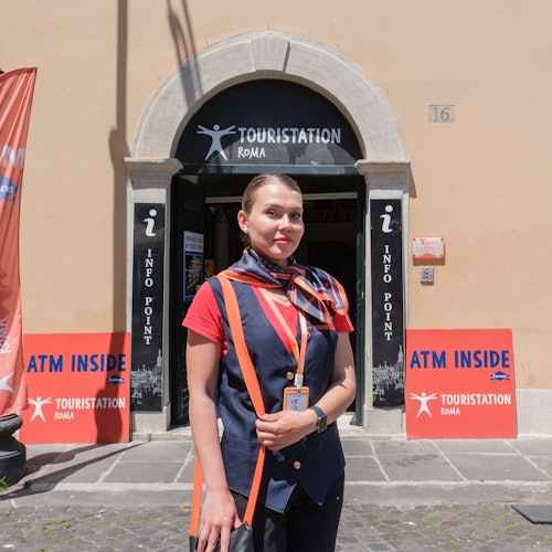 一位身穿制服的女士站在标有“Touristation Roma”的建筑前，建筑内有信息点和ATM的标识广告。