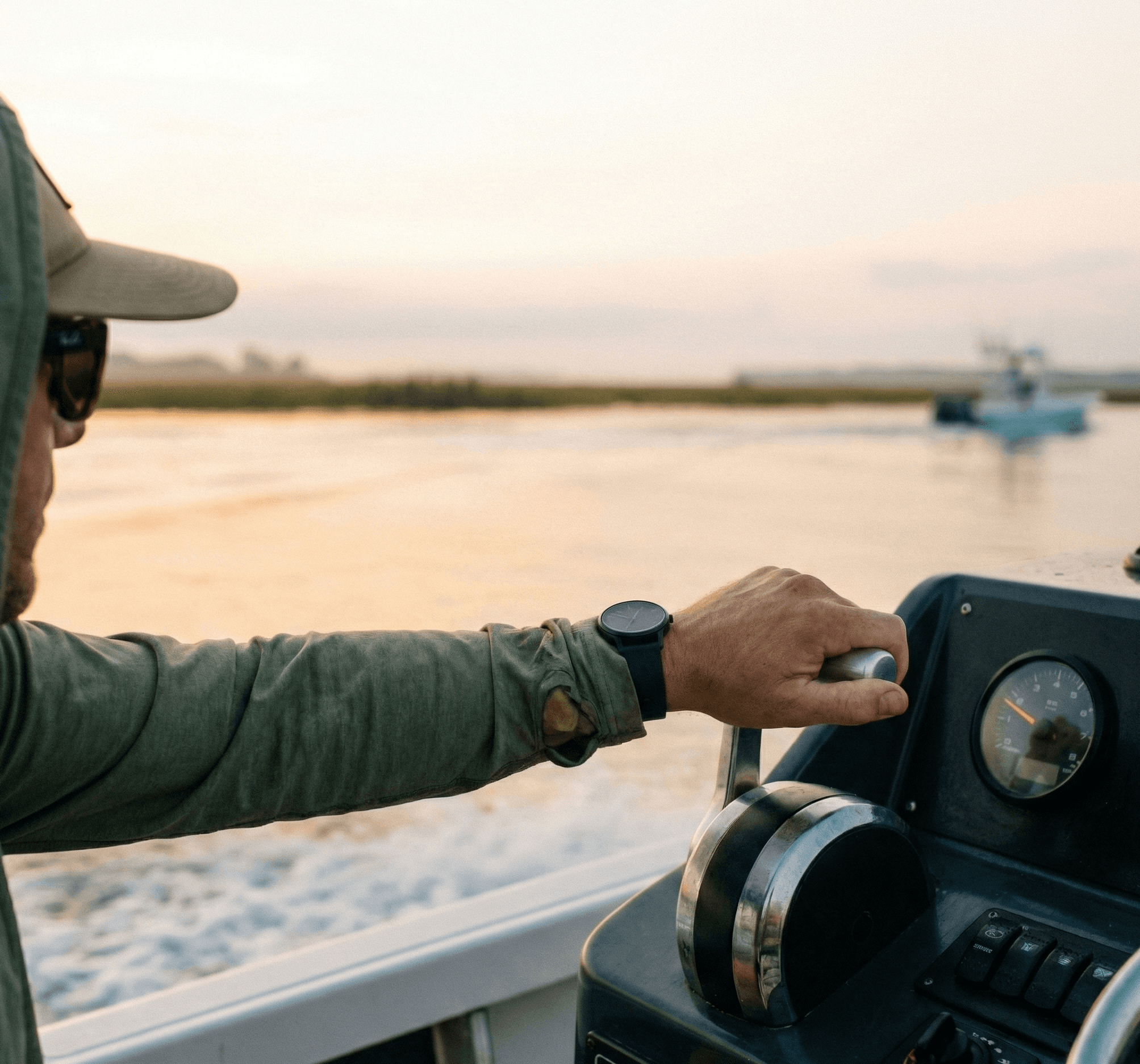 A close-up of a person’s hand gripping the throttle of a motorboat at sunrise, with calm water and a distant boat softly blurred in the background.