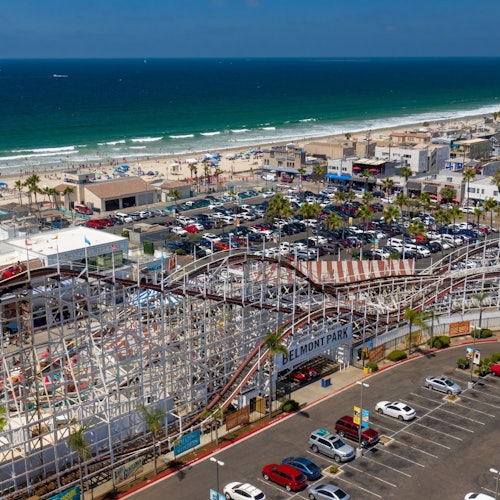 Una vista aérea de Belmont Park con una montaña rusa de madera, estacionamiento, bañistas y el océano de fondo.