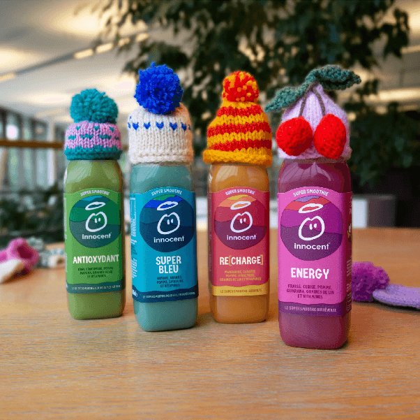 Quatre bouteilles de smoothies Innocent coiffées de petits bonnets tricotés, posées sur une table.