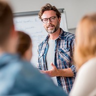 Lehrkraft steht im Zentrum eines Klassenzimmers und erklärt einer kleinen Gruppe von Schülern Lerninhalte, Whiteboard und Unterrichtsmaterialien im Hintergrund