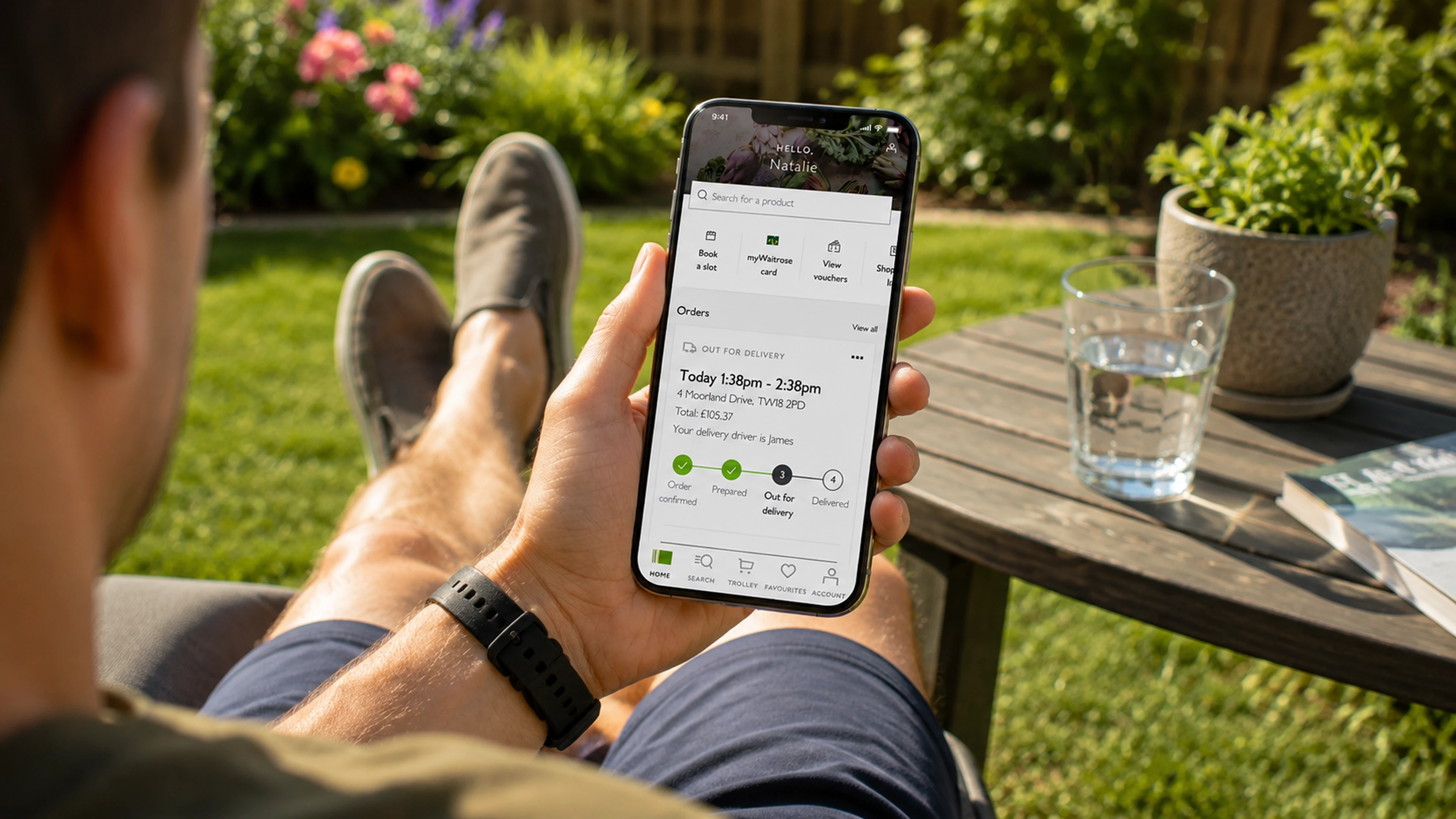 A man relaxing in a garden checking his phone, which shows the order status out for delivery.