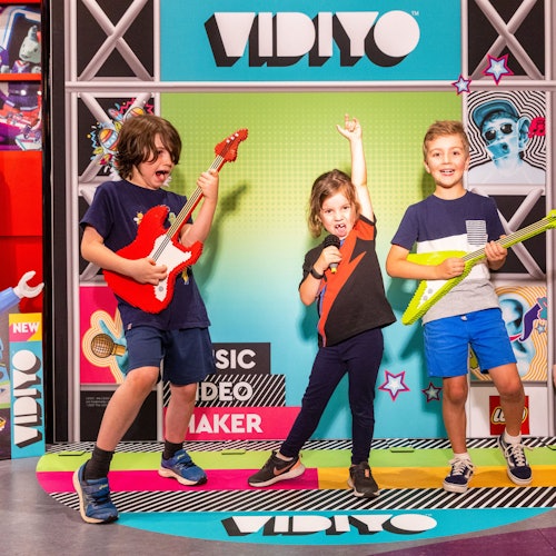 Troje djece pozira s igračkama gitarama i mikrofonom ispred šarene pozadine s riječju "VIDIYO."