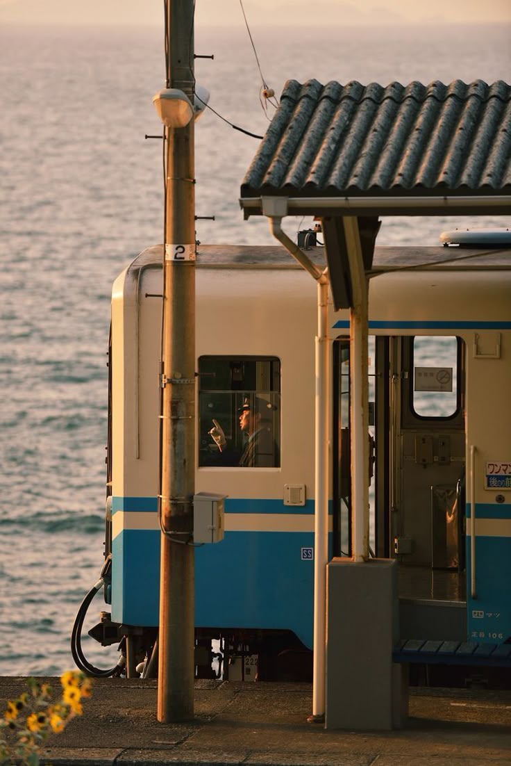 A train at a coastal station with a conductor visible in the cabin, surrounded by a serene sea backdrop and a pole adorned with greenery.