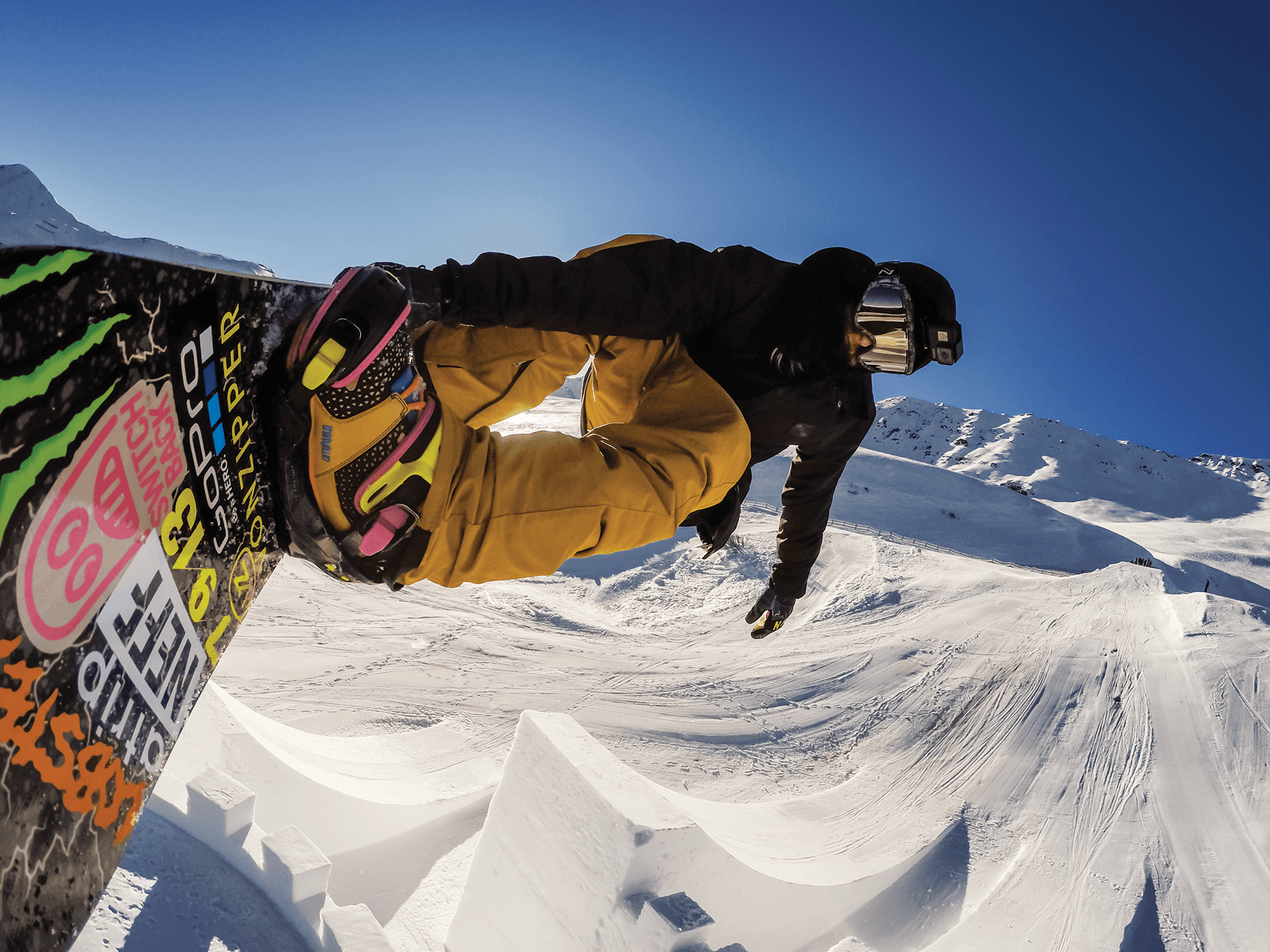 A GoPro camera mounted to the well-stickered board of professional snowboarder Halldor Helgason as he flies over a massive kicker built on a snow-covered Watles Ski Area in the Italian Alps. Halldor is wearing colorful boots and bindings, with tan pants, a black jacket, gloves, and beanie, with reflective snowboarding goggles and another GoPro on his head.