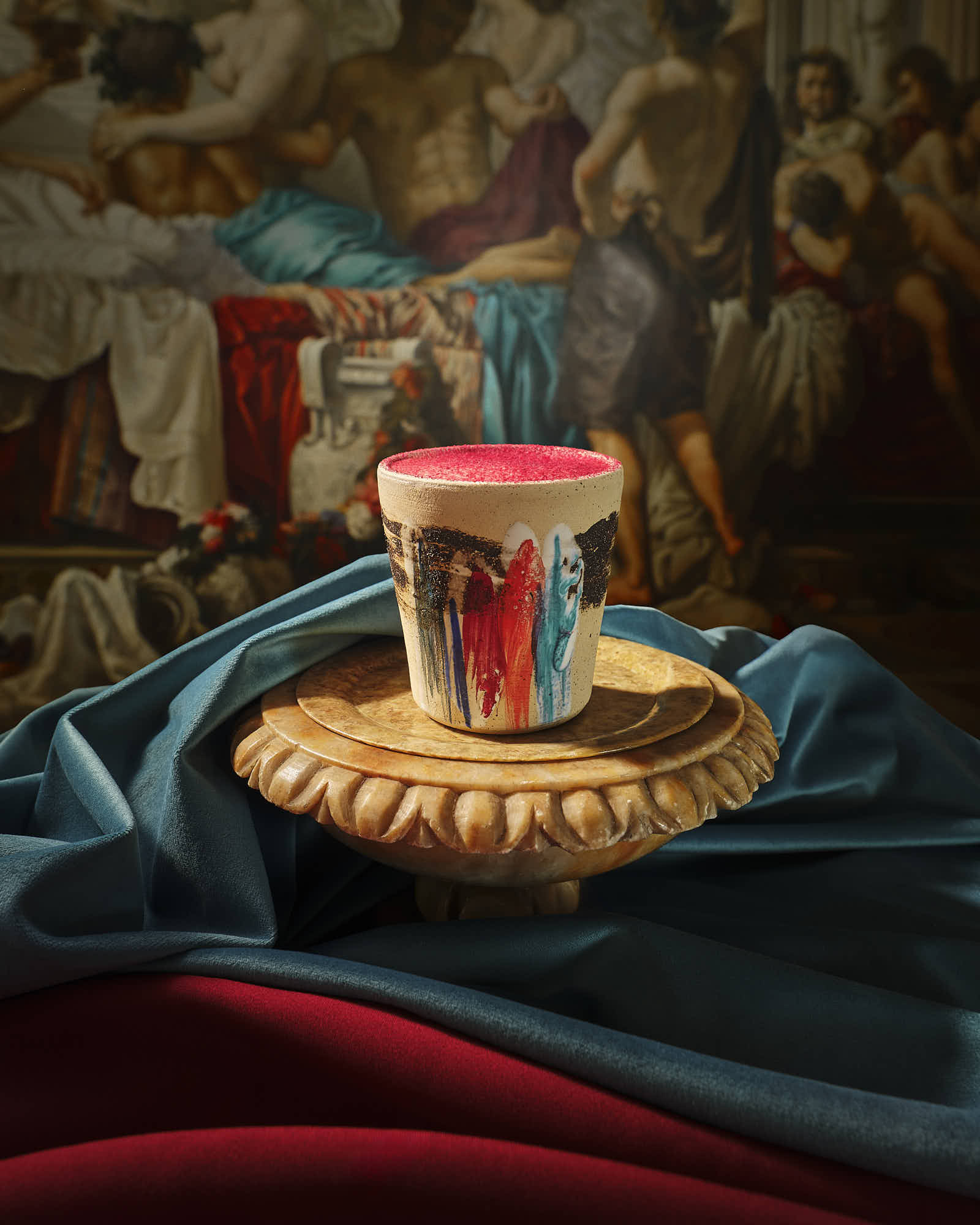 A decorative glass filled with a colorful drink sits atop a gold plate on textured fabric in rich colors.