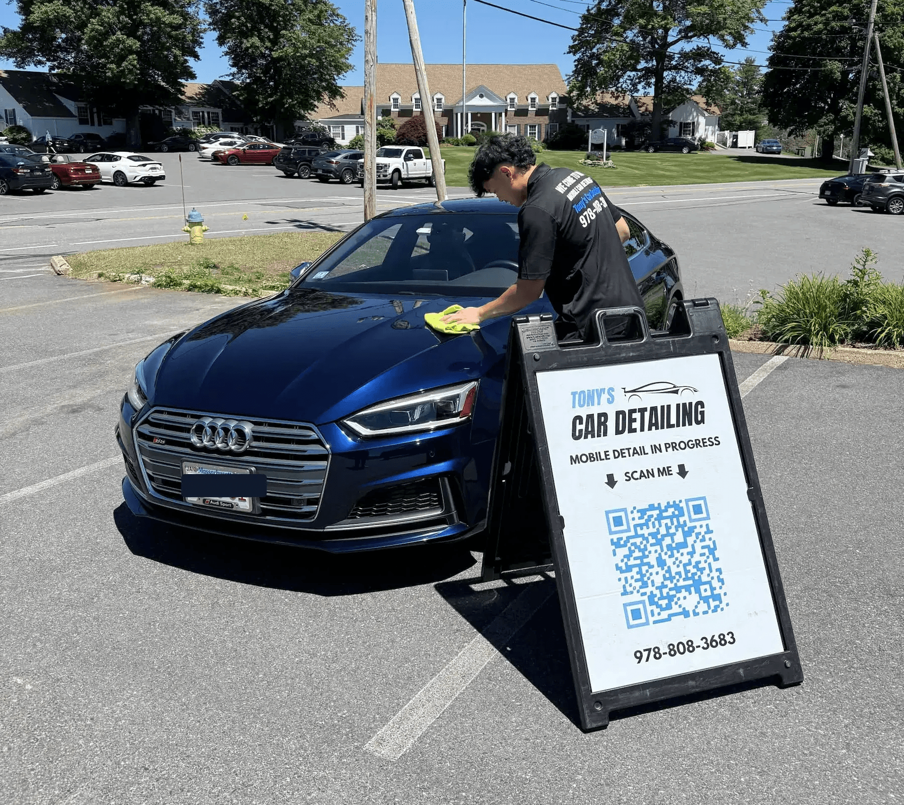 A mobile detailer wiping down the hood of a shiny, dark blue Audi sedan in a parking lot on a sunny day next to a "Tony's Car Detailing" A-frame sign with a QR code.