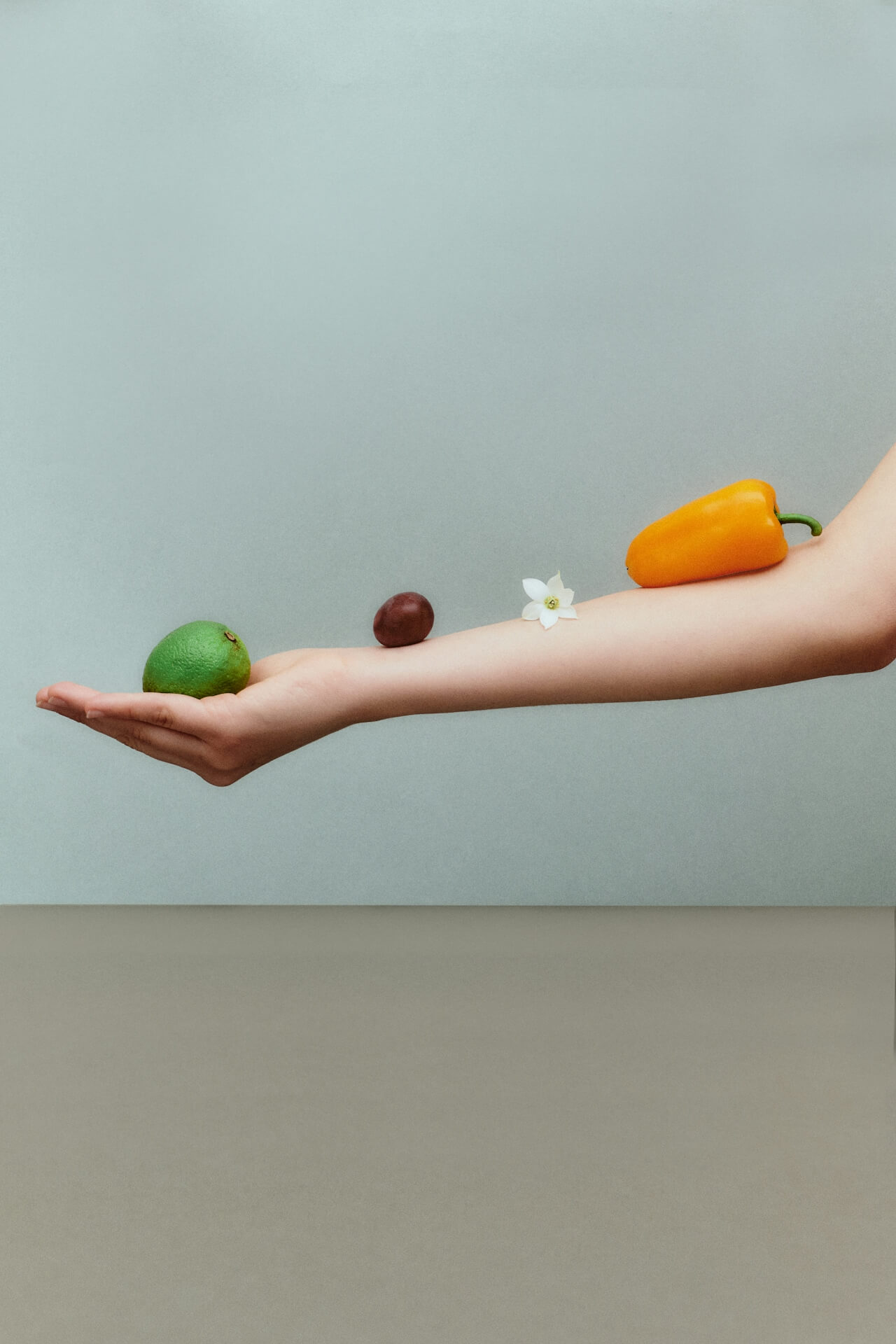 A girl's outstretched arm against a greyish-blue backdrop with an orange pepper, flower, grape and lime arranged across her arm