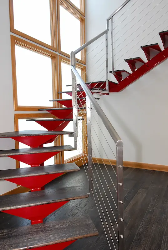 Portrait of a red, modern steel staircase with stainless steel handrails with cable rails. Large, 6 panel picture window with obscured glass, letting in abundant natural light. Irvine Transitional Modern Project. Photo by Chris Darnall.