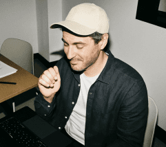 A person wearing a cap and a jacket smiles while sitting at a table, appearing engaged in conversation or thought.