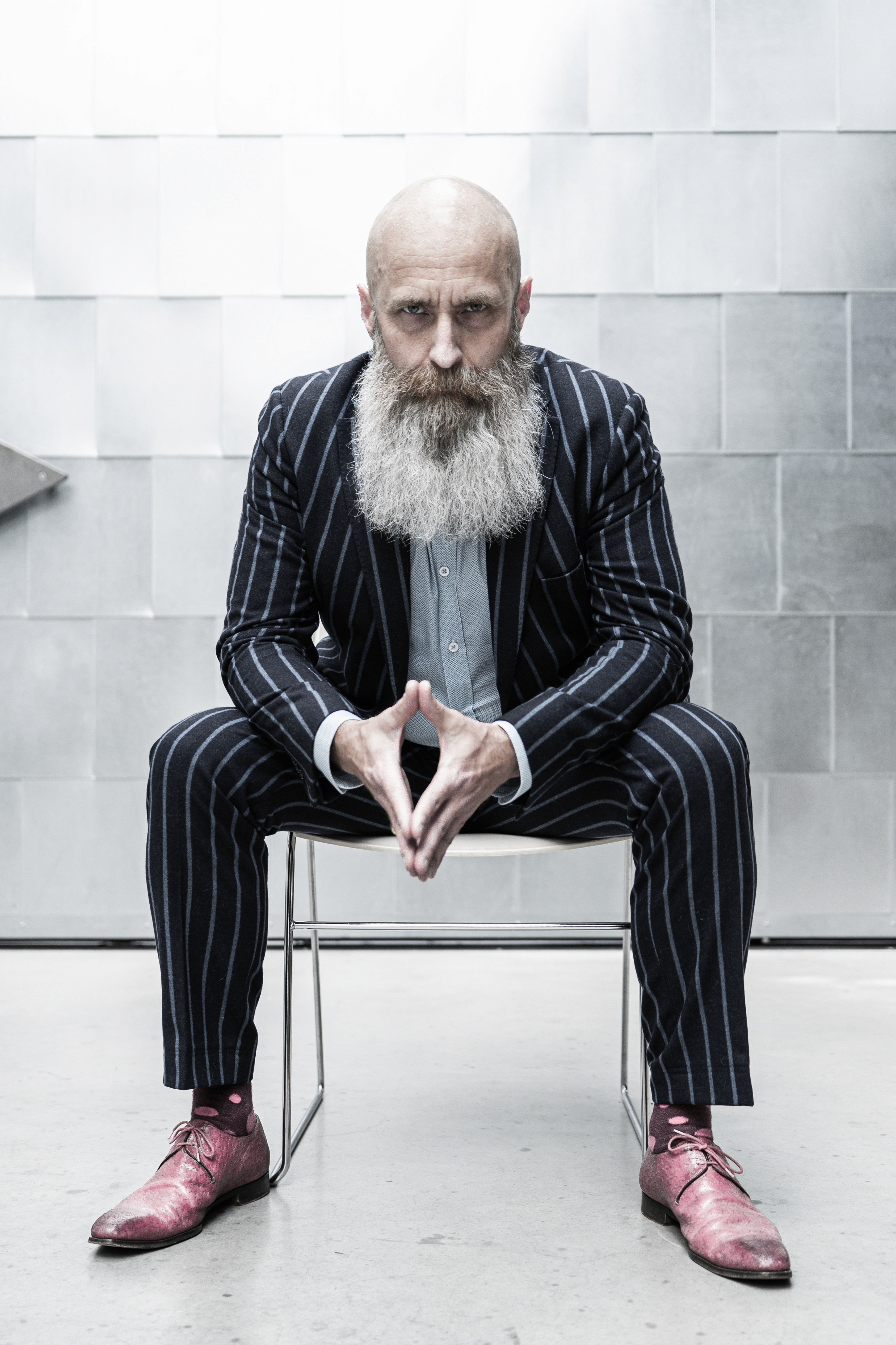 Bas Stronks, man met grijze baard en streepjespak en roze scheonen zittend in een industriele omgeving.
