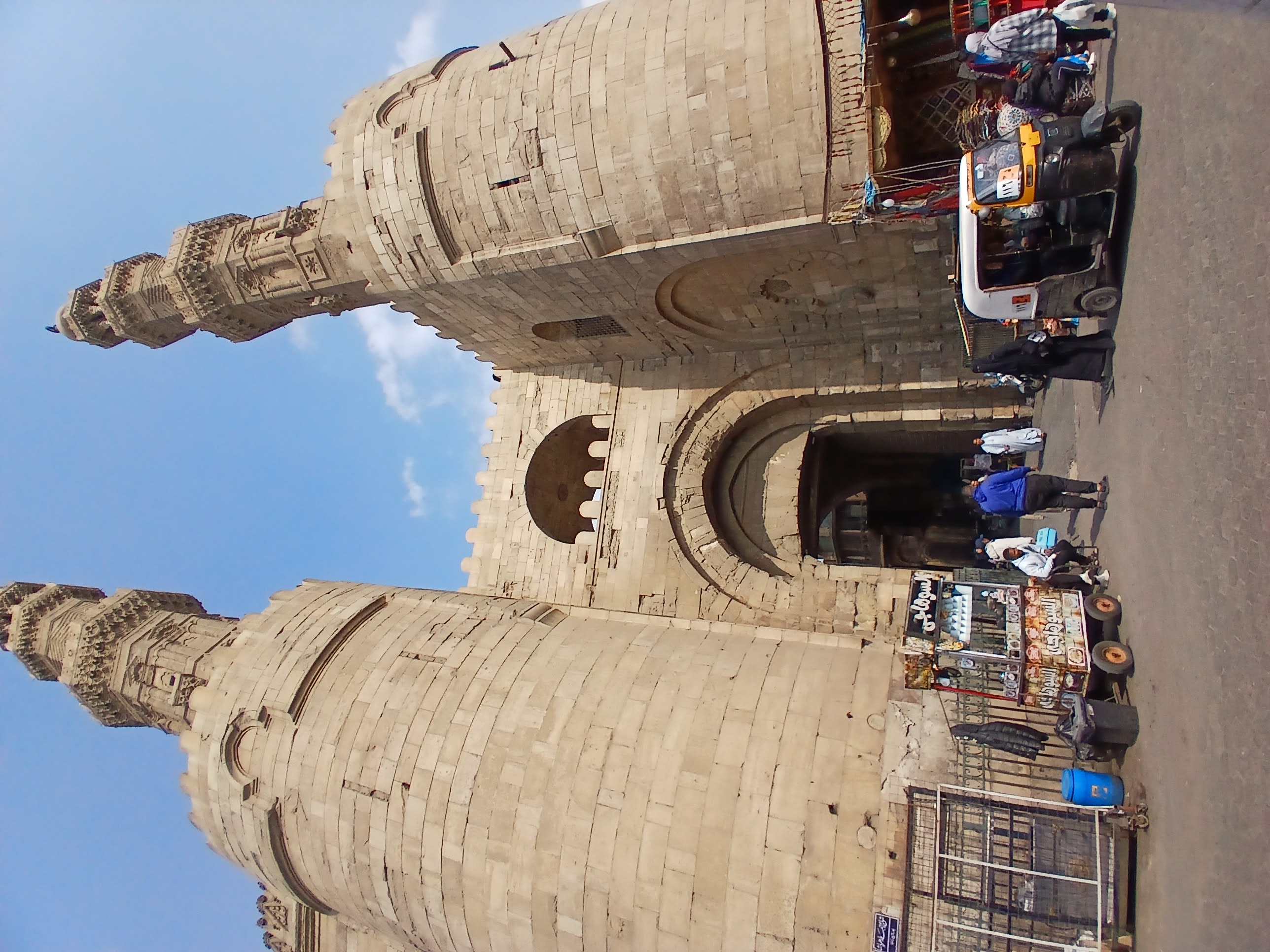 A gate from fatimi cairio, outside the gate are two large mnarets and some people and a rikshaw 