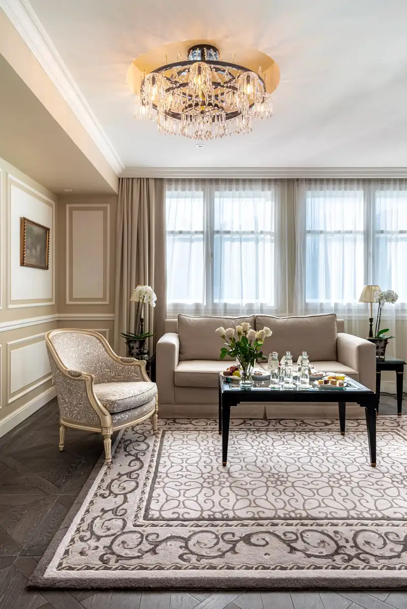 A formal living room with a beige sofa, an armchair, a chandelier, and a large patterned rug on a dark wood floor.
