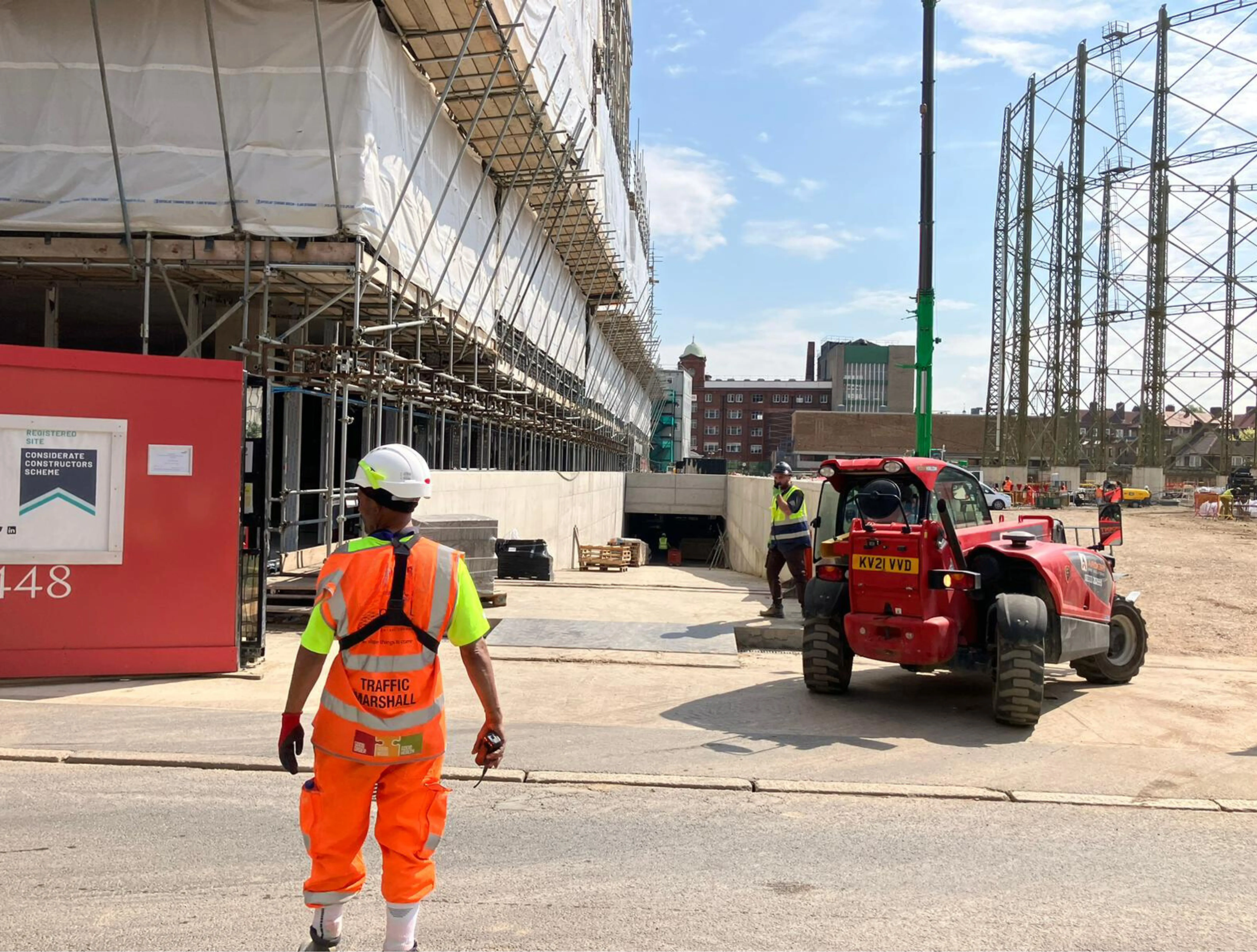 A construction worker in high-visibility gear stands near an urban construction site with partially completed scaffolding, while a red forklift operates alongside, set against a backdrop of clear blue skies and industrial structures.