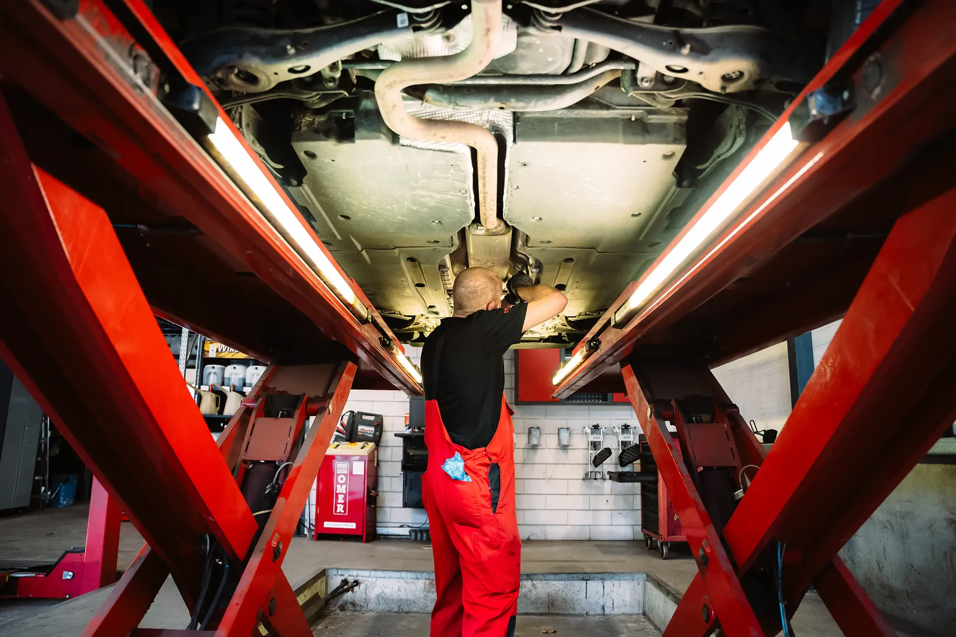 Inside the car dealership garage were a mechanic is looking at the bottom of a car while the car is on a car elevator