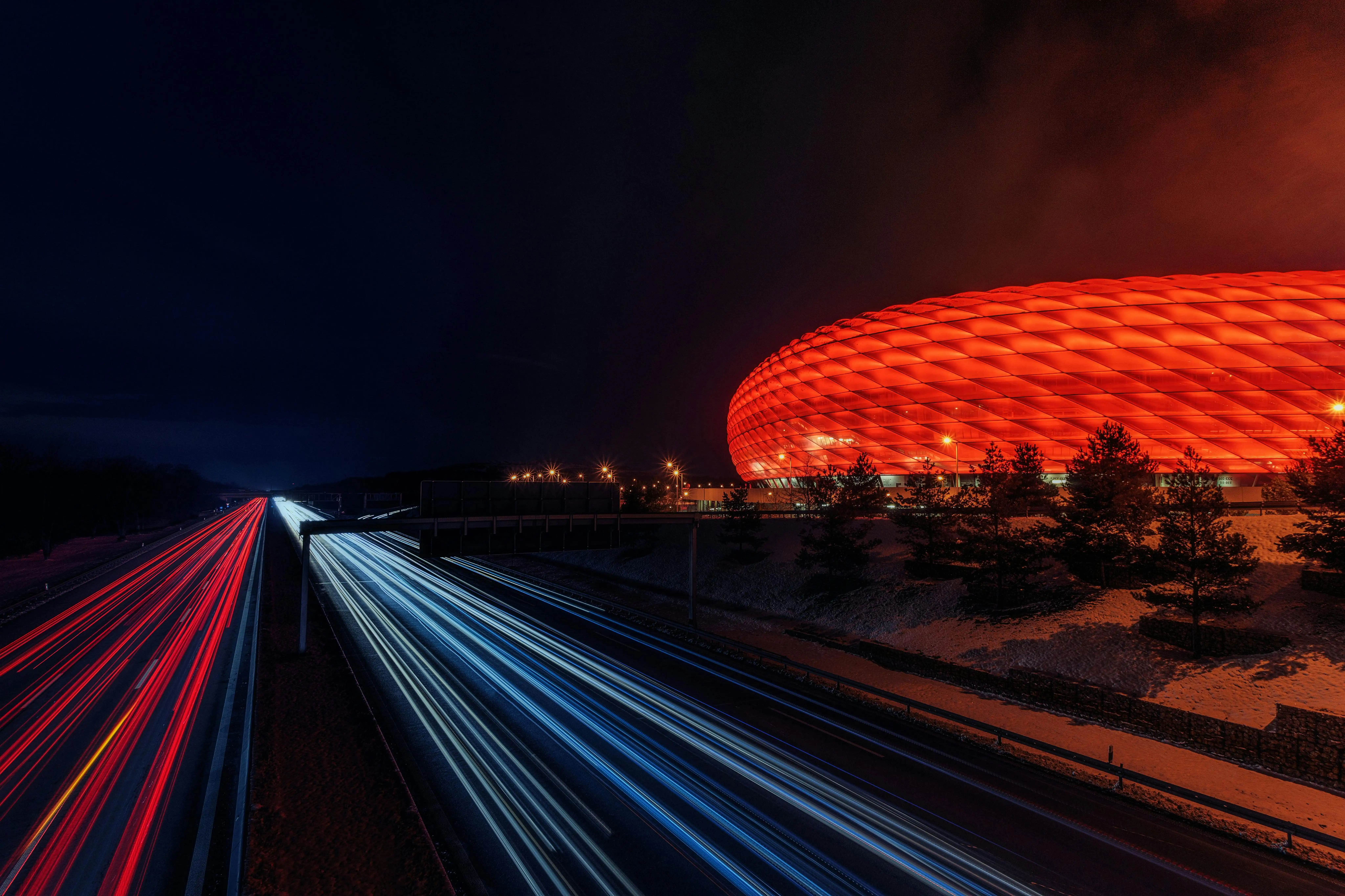 Illuminated modular stadium at night with dramatic LED facade and highway light trails, representing large-scale global event infrastructure.