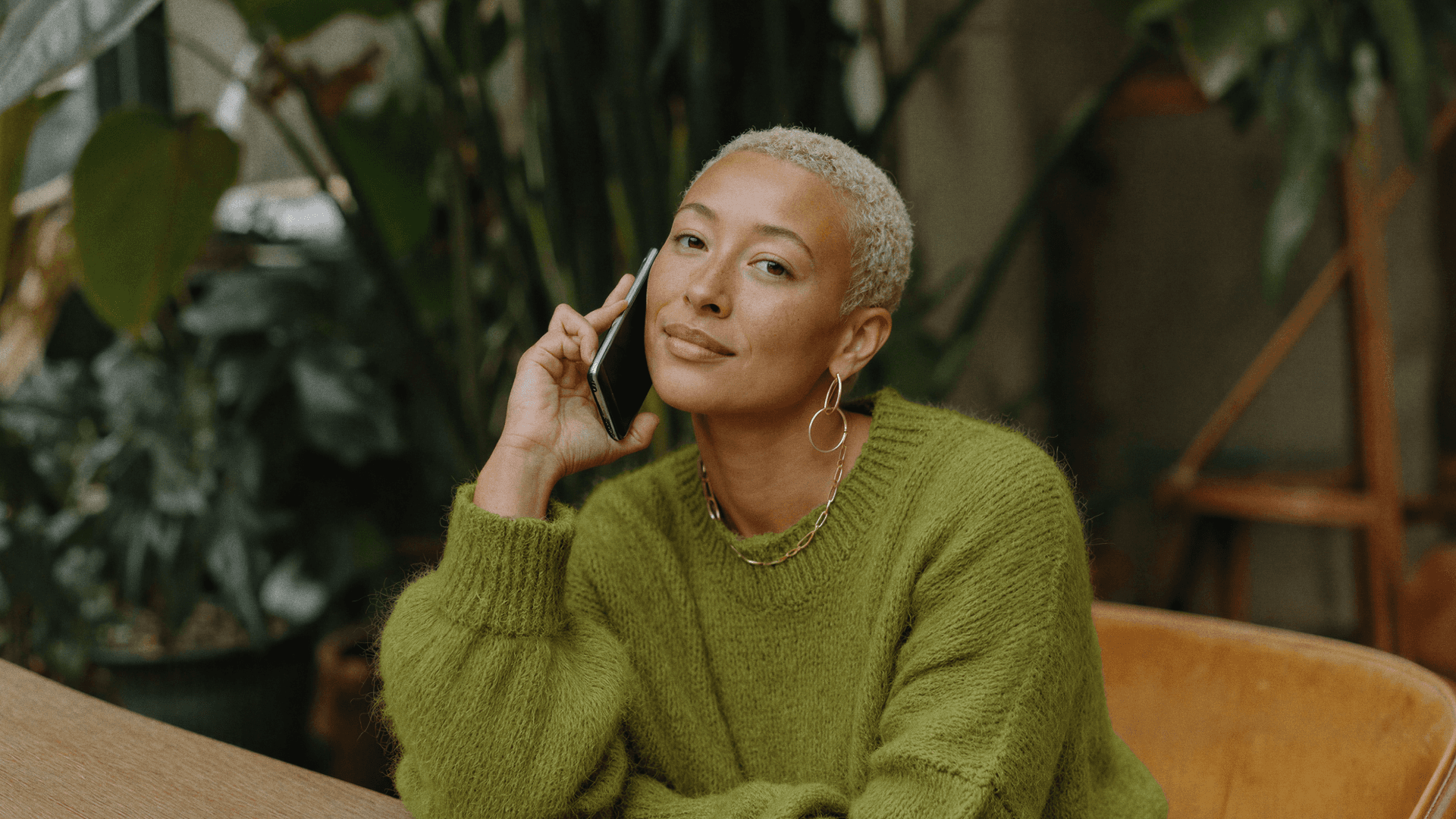Mulher sentada em cadeira de madeira, com celular no ouvido e expressão serena, com folhas e vasos ao fundo de uma clínica automatizada pela Tivita