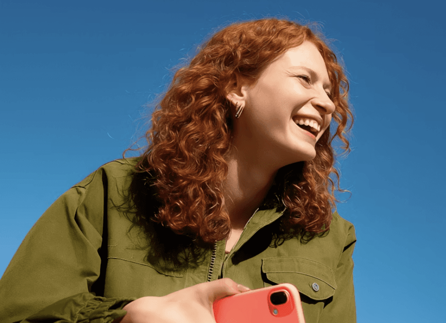 A cheerful person with blonde hair and a purple sweater enjoys using their green smartphone under a clear blue sky.