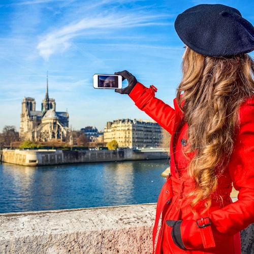 Person in a red coat and black beret taking a photo with a smartphone, capturing a riverside landmark under a blue sky.