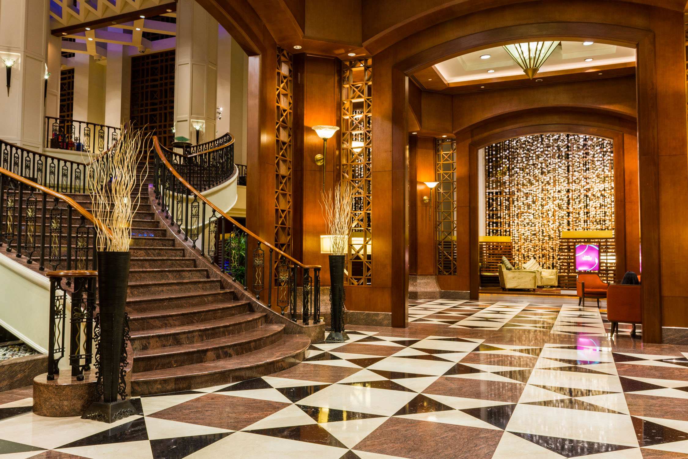 A grand hotel lobby with a wide staircase, ornate gold railings, and a checkered marble floor under warm lighting.