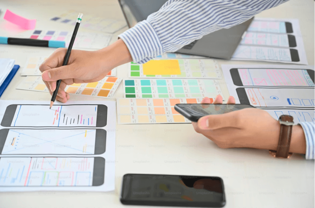 Designer sketches mobile app wireframes on paper while holding a smartphone, with color palettes and interface layouts spread across the desk.