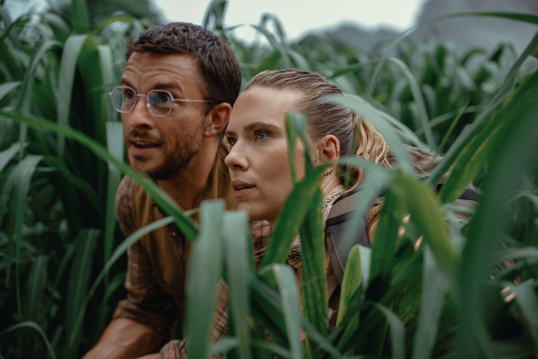 Jonathan Bailey and Scarlett Johansson in 'Jurassic World', crouching in tall grass with fascinated expressions.