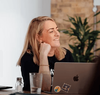 Woman in a tailored beige suit holding a structured brown leather handbag, standing in front of a softly lit modern background.