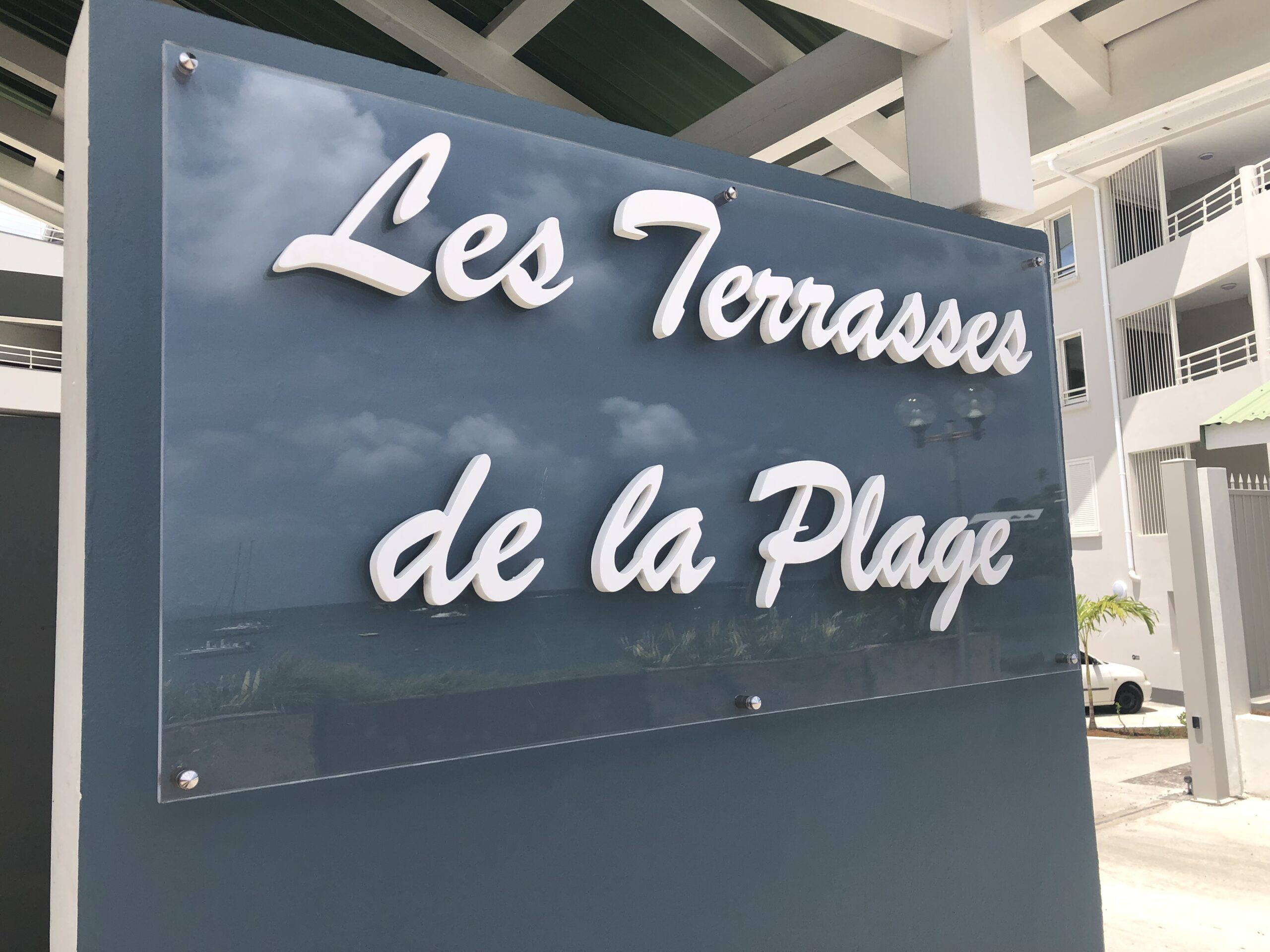 Enseigne portant l'inscription « Les Terrasses de la Plage » en lettres blanches élégantes sur fond bleu réfléchissant, fixée à la façade d'un bâtiment moderne.