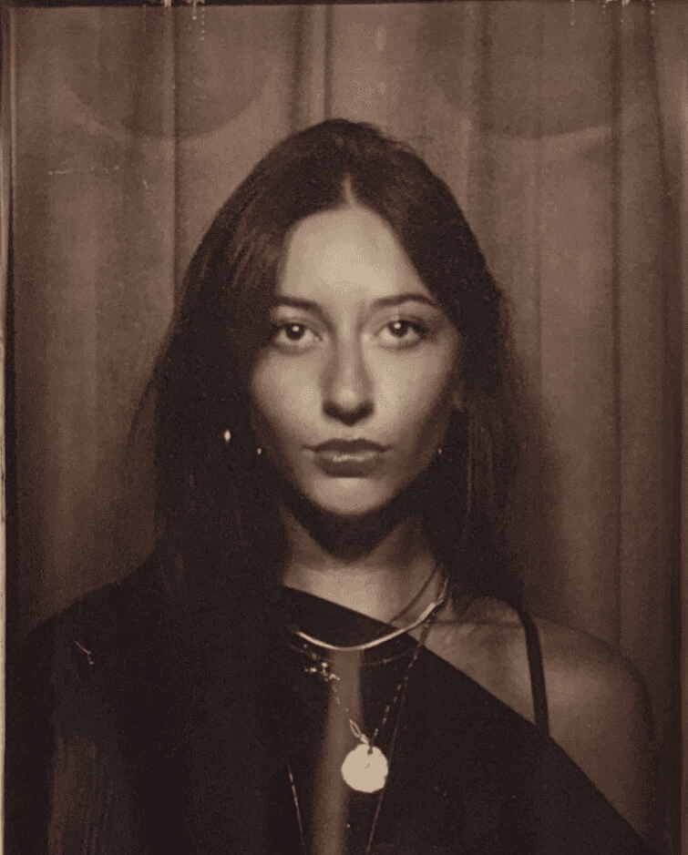 A photobooth still of a young woman with straight dark hair outing while weating an off the shoulder black top and a pendant neckalce