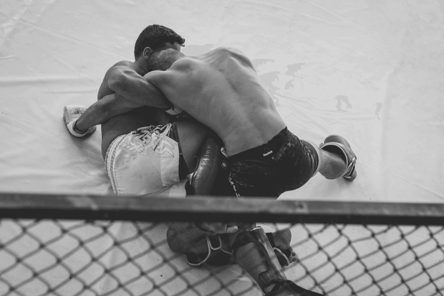 Two MMA fighters grappling on the mat near the cage wall during training.