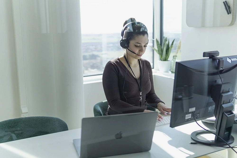 A young person with curly hair smiles at the camera while sitting in front of a laptop against a dark background.