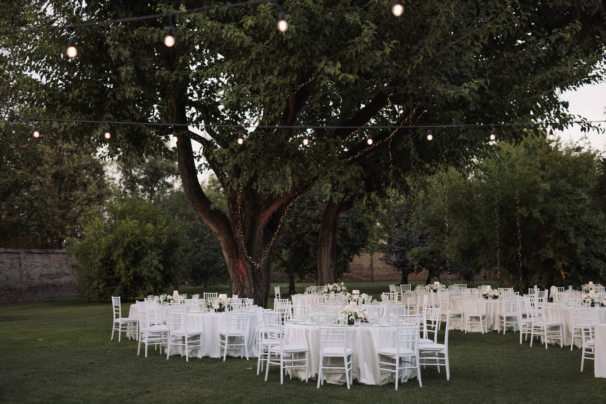 Tavoli rotondi con sedie bianche sotto il grande albero storico di Villa Negri, allestimento matrimonio in giardino