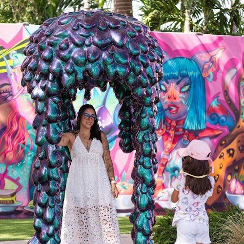 一位穿着白色连衣裙的女士站在一个紫色和绿色雕塑下方，而一个戴粉色帽子的孩子在一旁观看。背景是色彩斑斓的壁画。