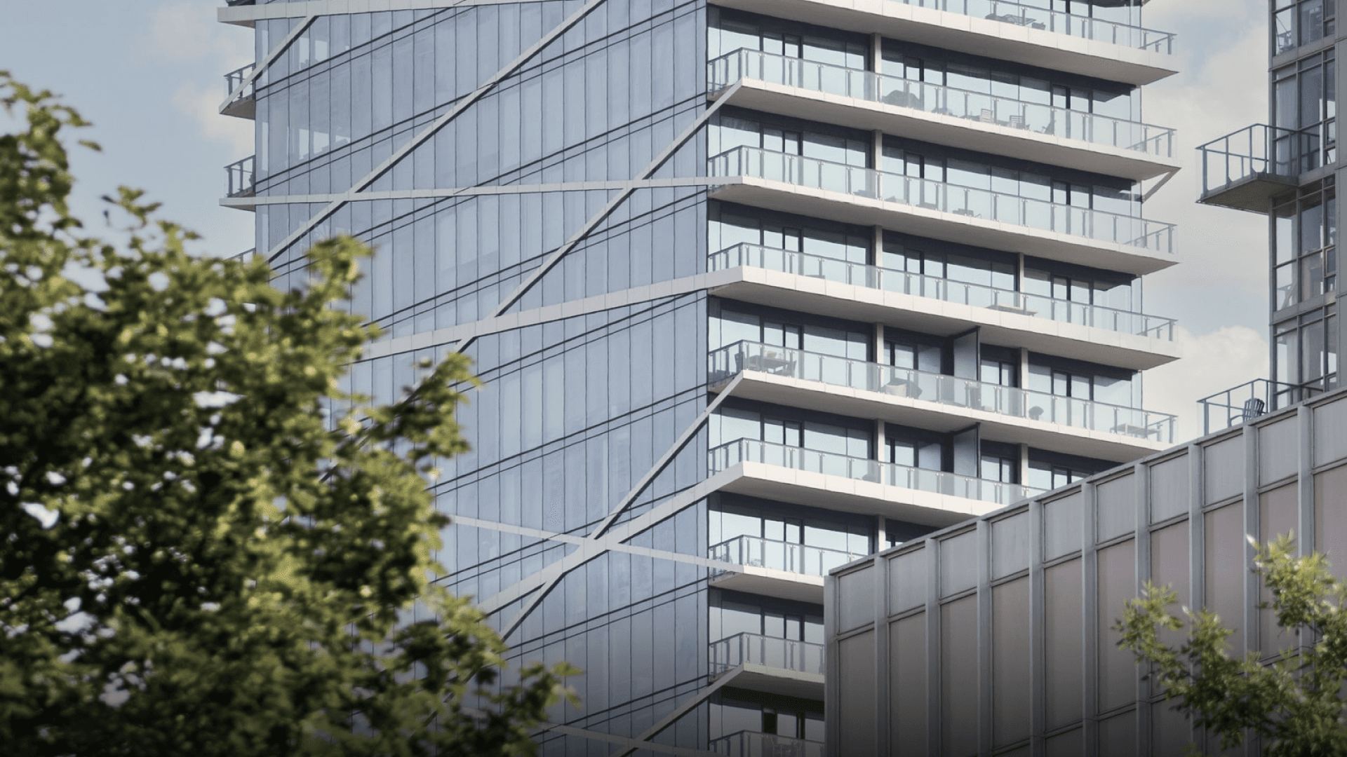 Close-up of a modern high-rise building featuring a geometric design with diagonal lines across its glass facade. The building includes rows of balconies with glass railings, creating a clean, contemporary aesthetic. In the foreground, partially blurred green foliage adds a natural contrast to the urban architecture.