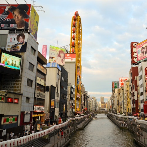 在繁华的城市环境中，城市运河两侧排满建筑物，色彩斑斓的广告牌，还有一个独特的黄色摩天轮。