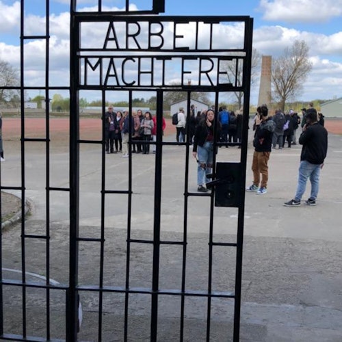 사람들 한 무리가 "ARBEIT MACHT FREI"라는 글귀가 새겨진 철문 뒤에 서 있습니다. 하늘은 부분적으로 구름이 끼어 있습니다.