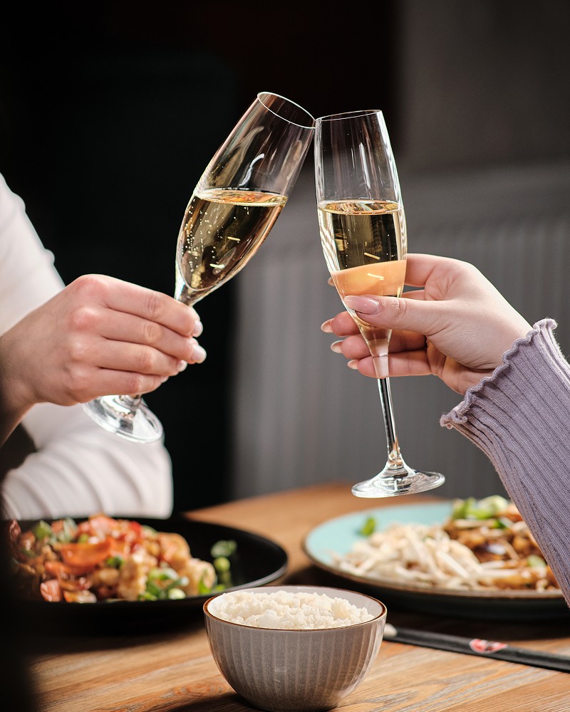 Fotografia kulinarna przedstawiająca zbliżenie na dwie osoby stukające się kieliszkami szampana przy kolacji